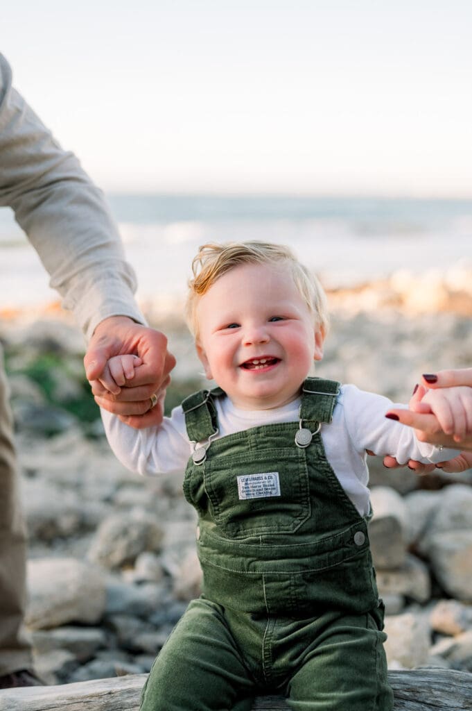 A little boy in green overalls smiles for the camera on a rocky beach.