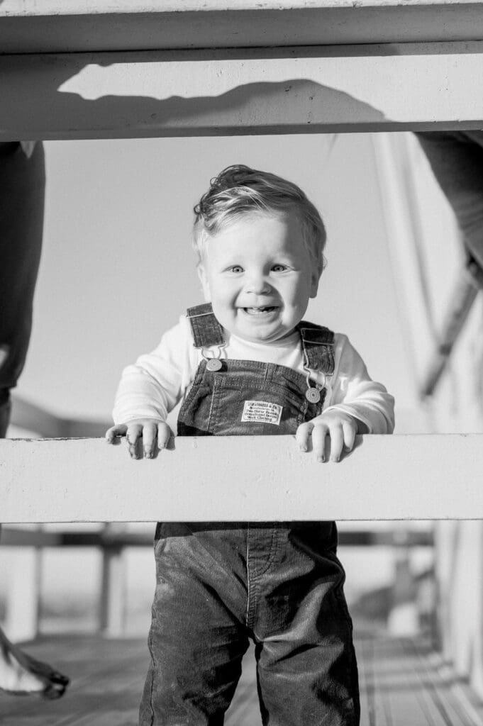 A black and white photo of a little boy on a lifeguard tower smiling.
