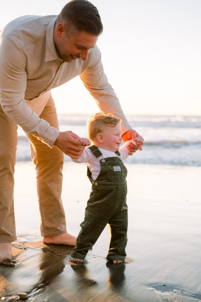 A dad holds his young son's hands as he tries to walk on a golden beach at sunset.