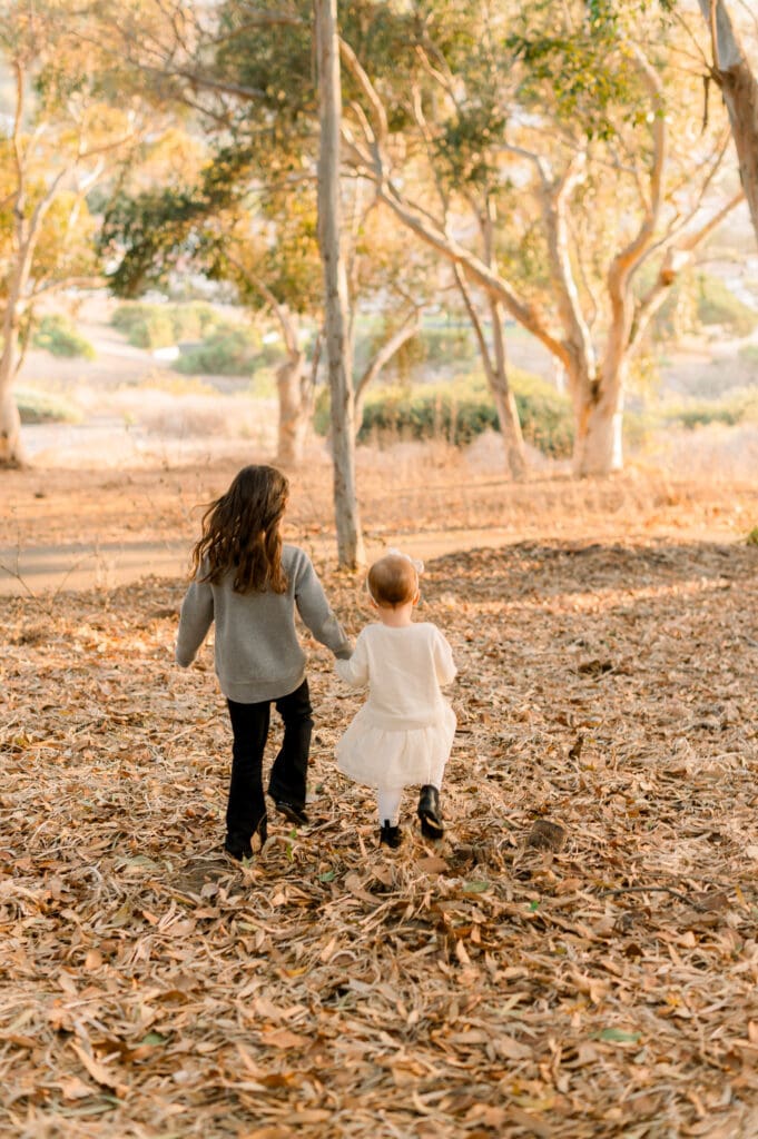 Two sisters run hand in hand through a wooded area.