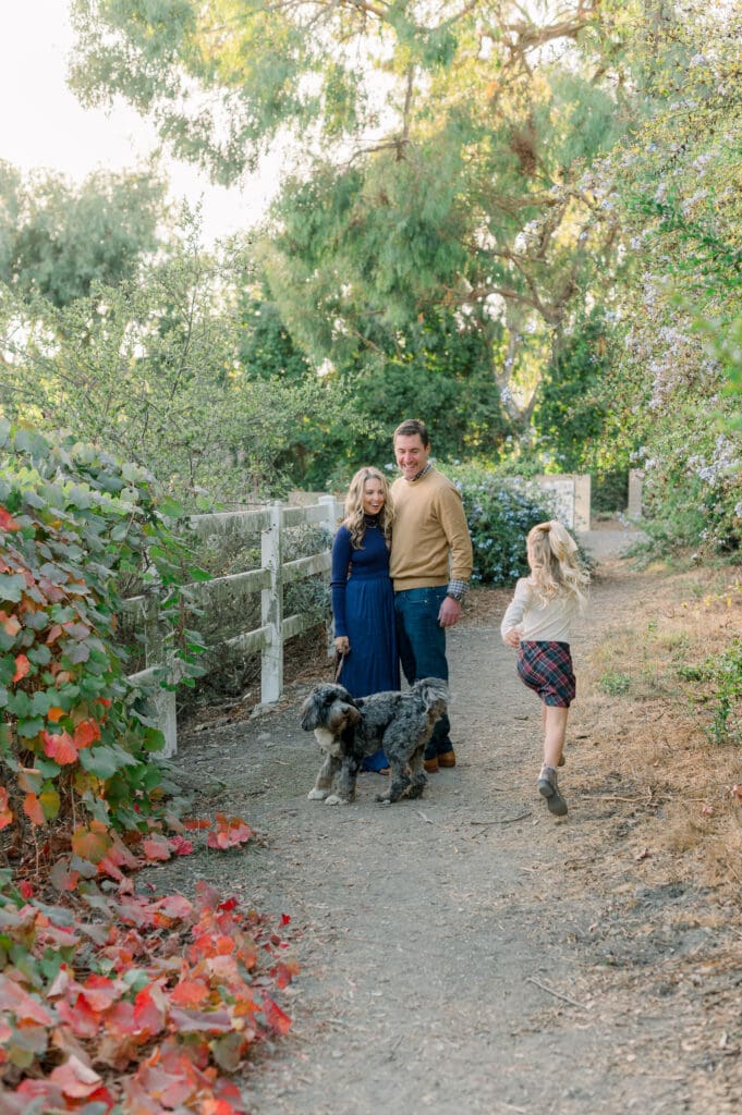 A mom, dad, and young girl taking fall family photos with their dog on a beautiful trail.