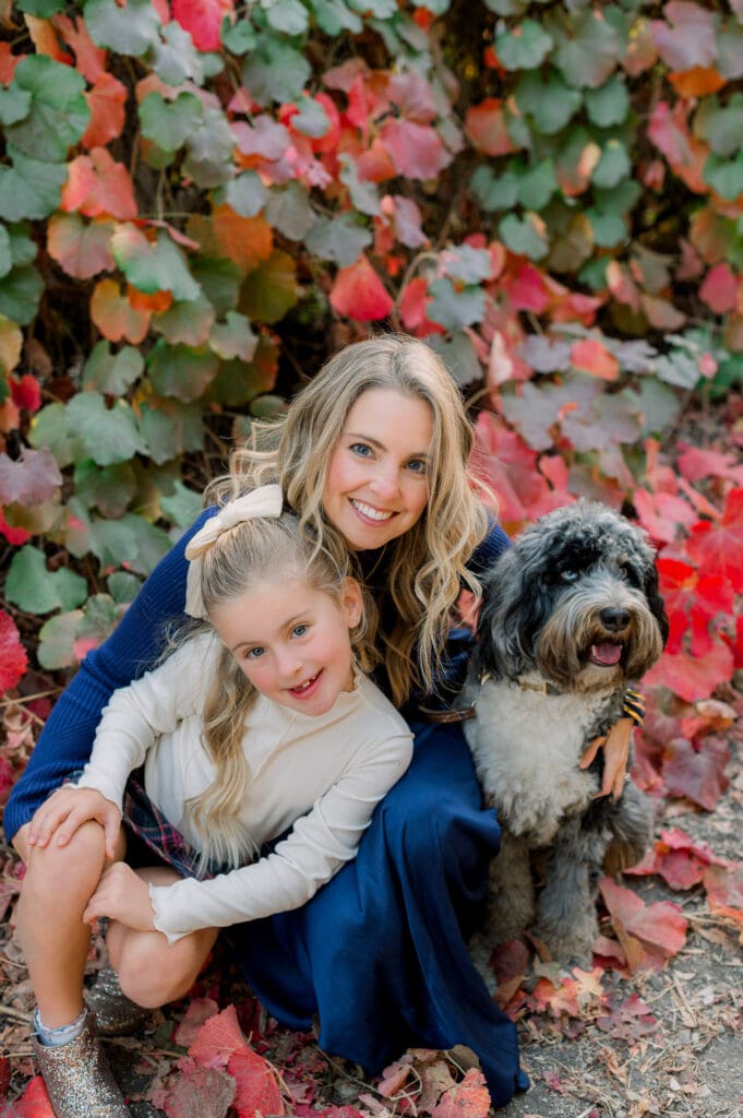 A photo of a mom daughter, and dog smiling at the camera in front of red and green leaves.