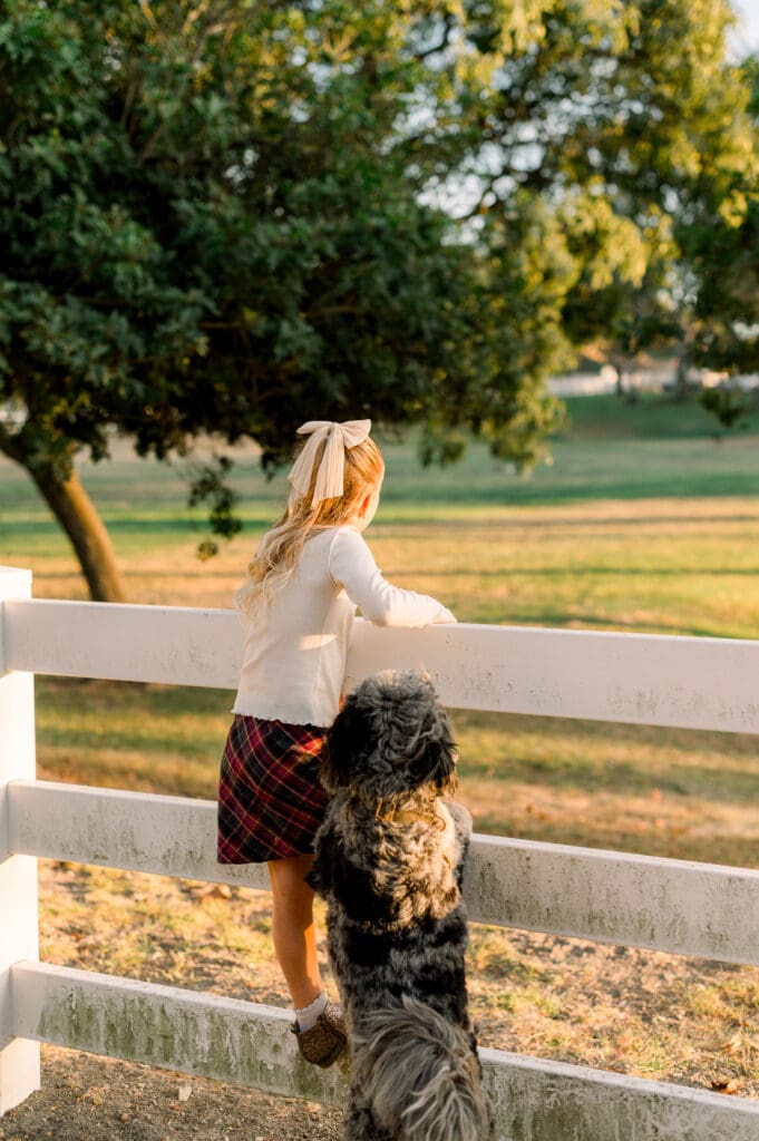 A little girl climbing on a white fence while her dog looks on.