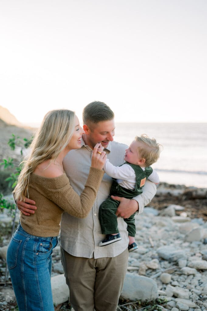 A mom, dad, and little boy laugh together on the beach during a fall family photoshoot.