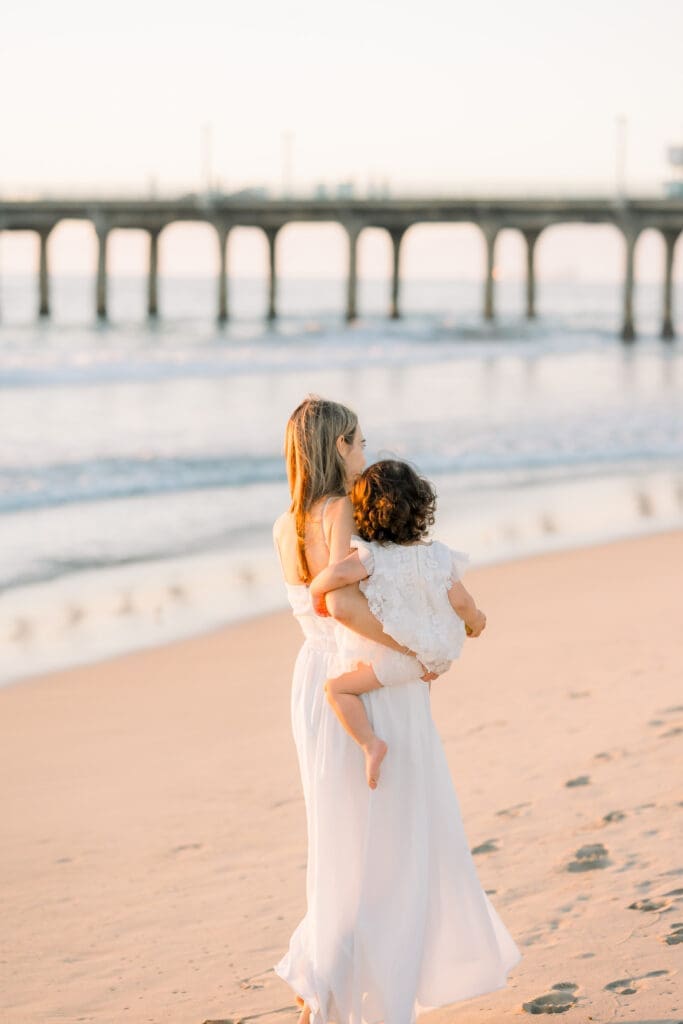 A mom holding her young daughter and walking on the beach as a Manhattan Beach family photographer takes their photo.