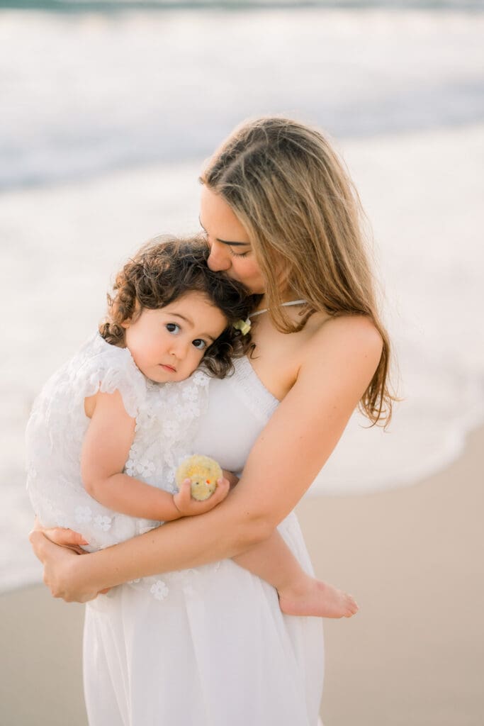 Mom kisses her daughter's hair while taking family photos on the beach.