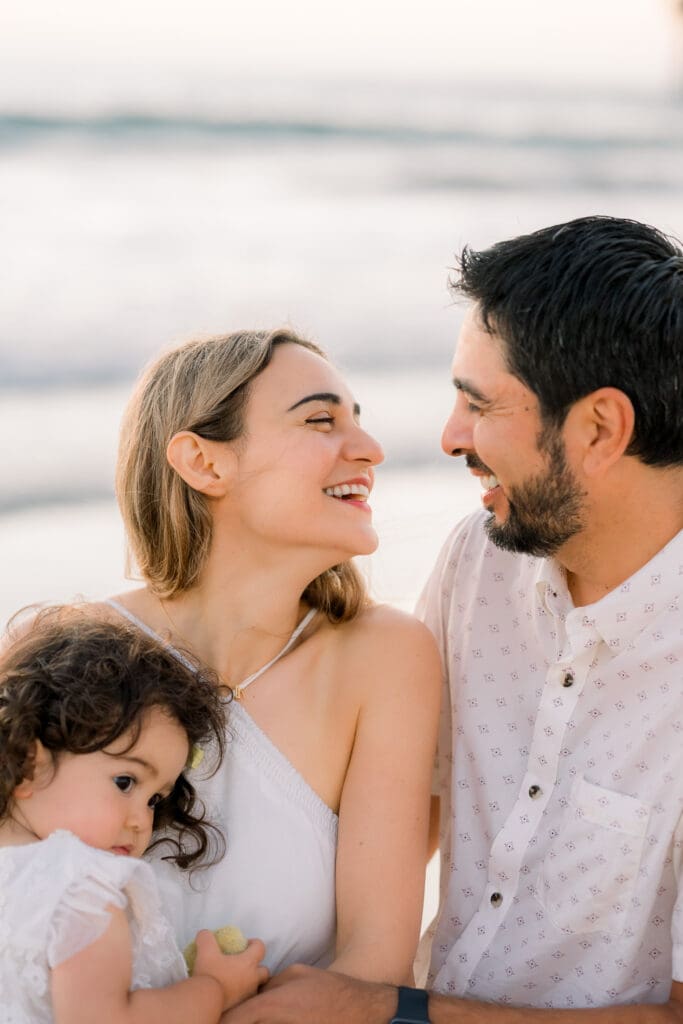 A Manhattan Beach family photographer takes a photo of a couple laughing on the beach as they hold their little girl close.