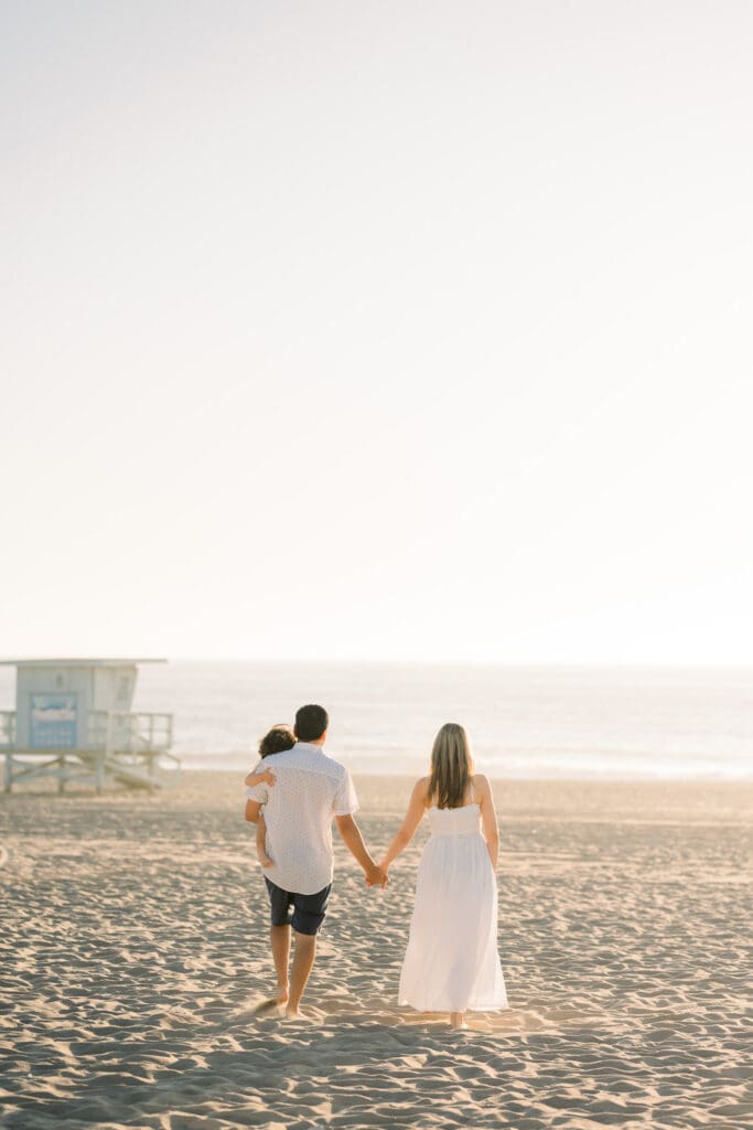 Mom and dad hold hands and walk on the beach with their baby during a family photography session in Manhattan Beach.