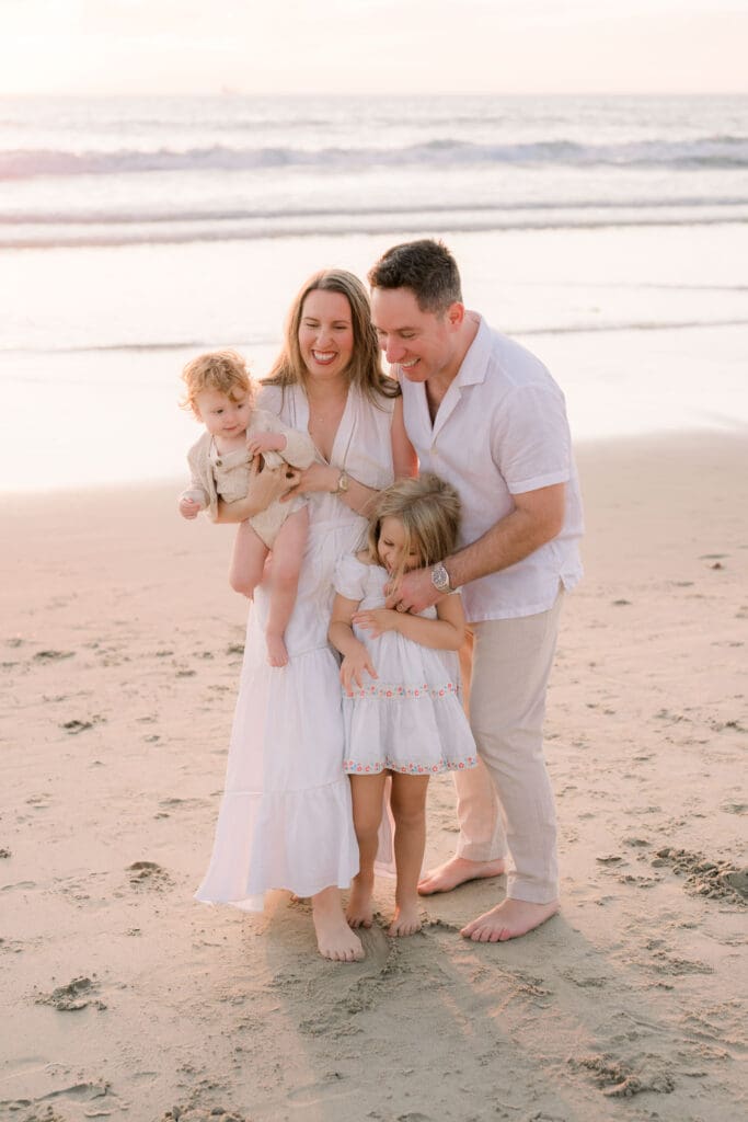 A family snuggling on a beach while taking family photos on the beach.  They are wearing all white clothing.