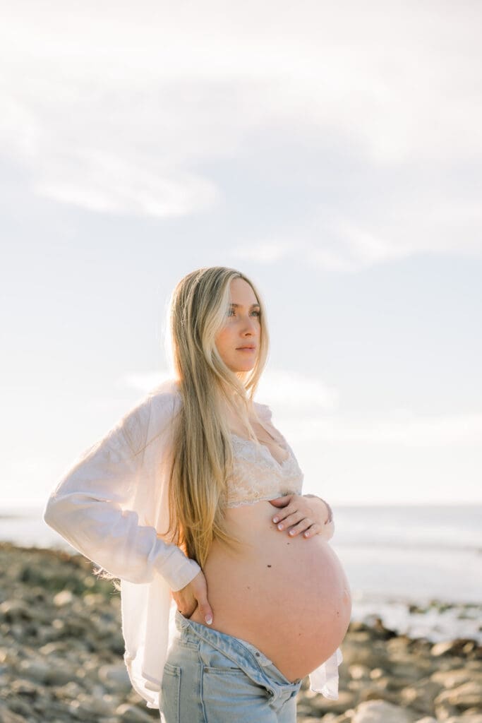 A pregnant woman wearing jeans and a white linen shirt poses for maternity photos on a South Bay beach.