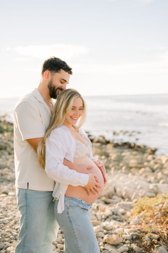 A couple posing for maternity photos at the beach in jeans and white shirts.