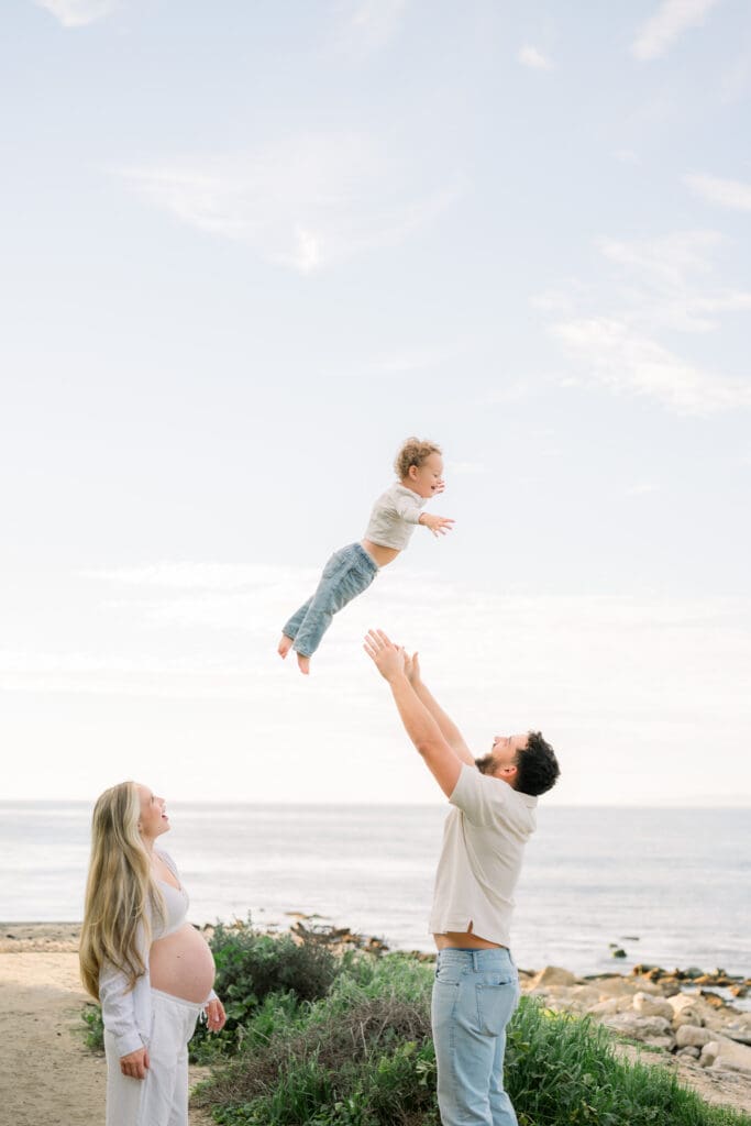 A casual beach maternity photo of a dad throwing his son in the air while the pregnant mom watches.