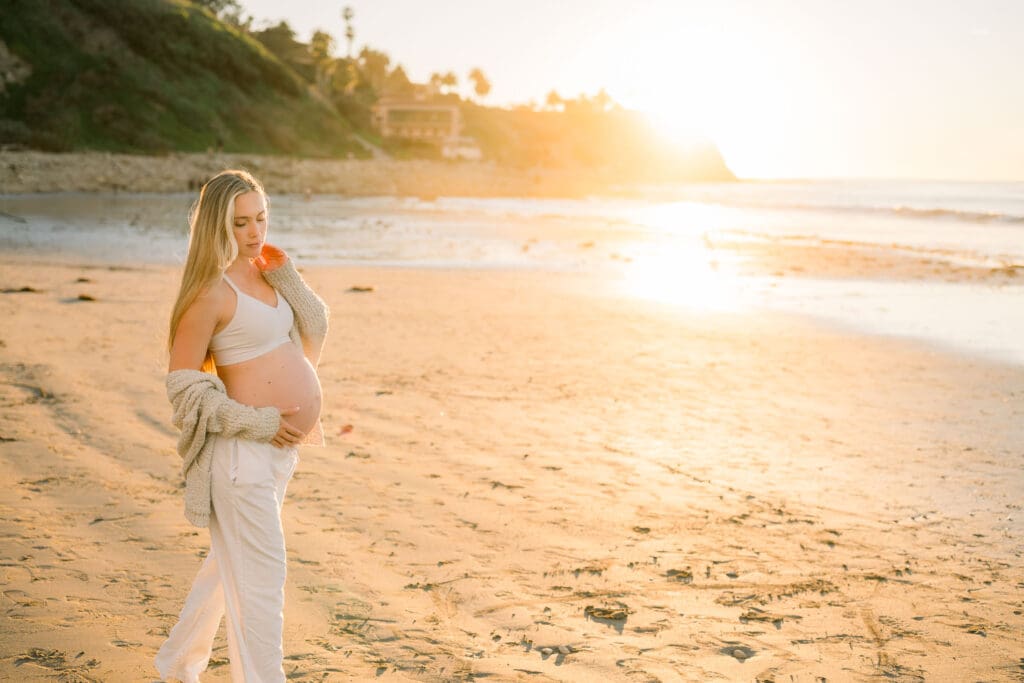 A pregnant woman walking on the beach at golden hour sunset.