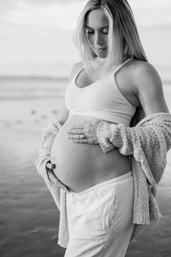 A black and white photo of a pregnant mother to be wearing white pants and a sports bra while holding her bare belly.