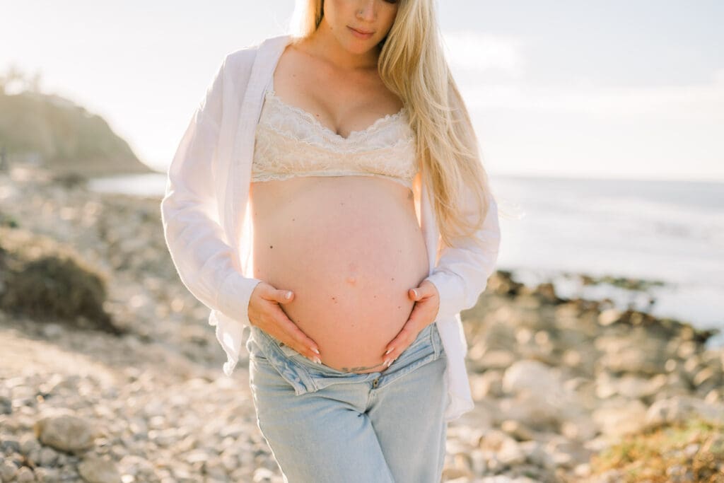 A pregnant woman holds her belly while taking casual beach maternity photos in the South Bay.