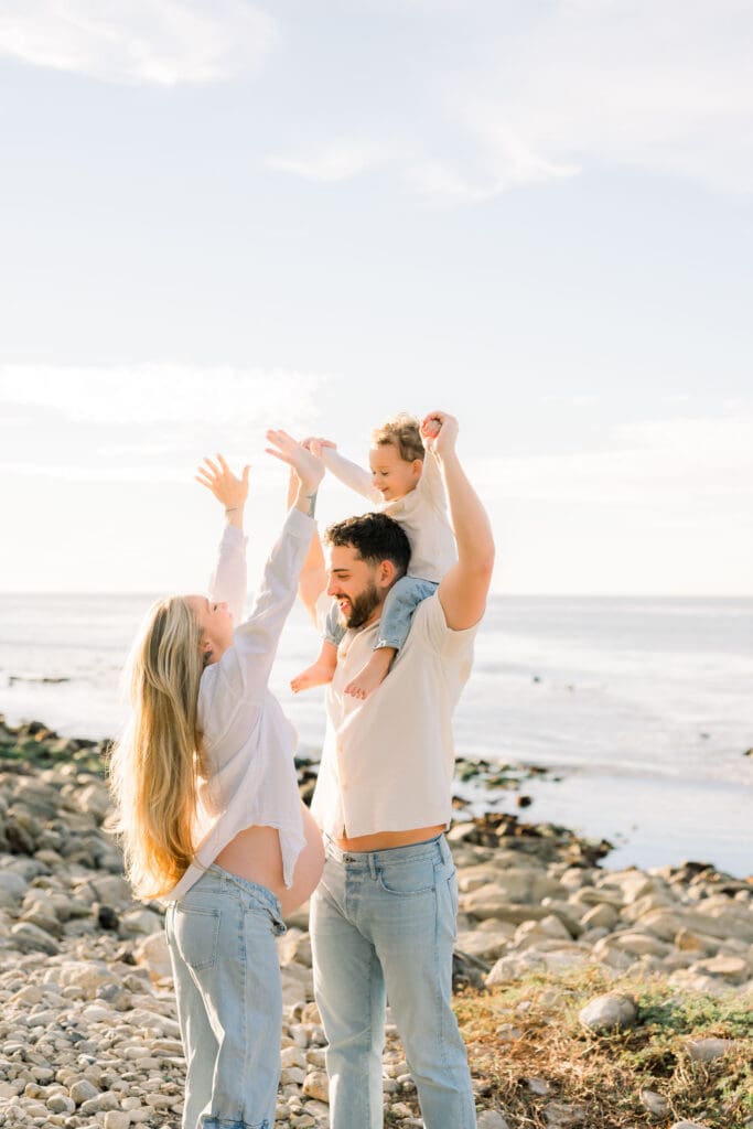 A little boy on his dad's shoulders laughing with his pregnant mom on the beach.