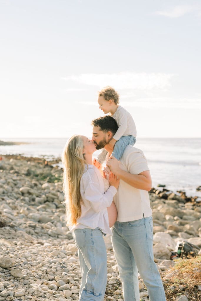 A little boy on his dad's shoulders while the dad kisses the pregnant mom.