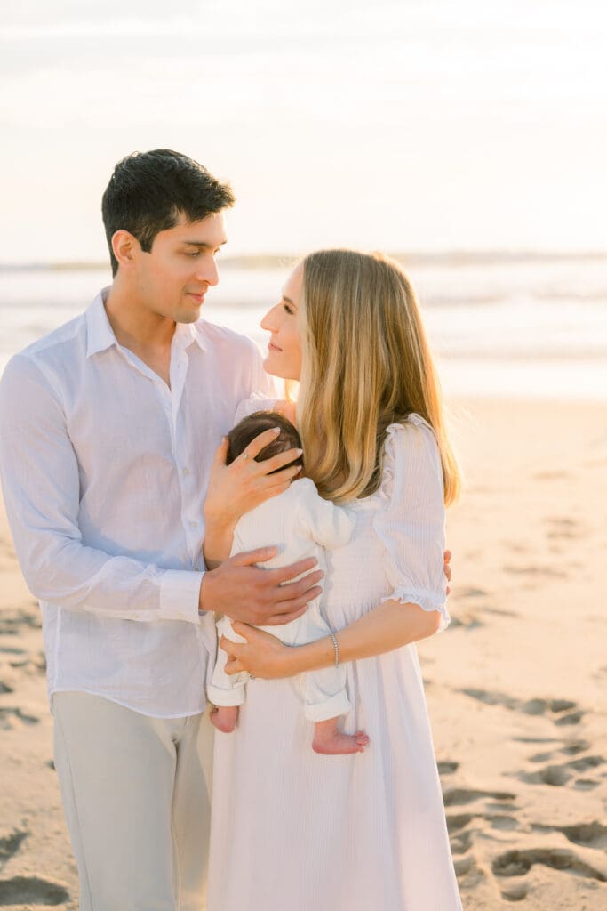 Dad and mom look at each other lovingly while holding their newborn baby on the beach at sunset.