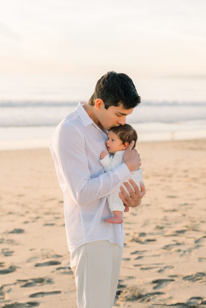 Dad kisses his newborn baby while taking newborn photos on the beach in Hermosa Beach.