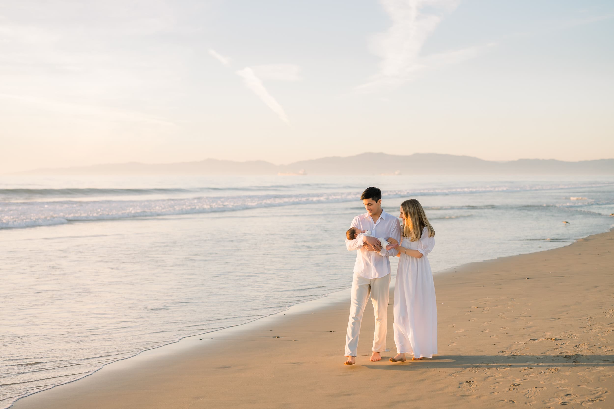 A Hermosa Beach newborn photographer captures photos of new family walking on the beach with their baby at sunset.