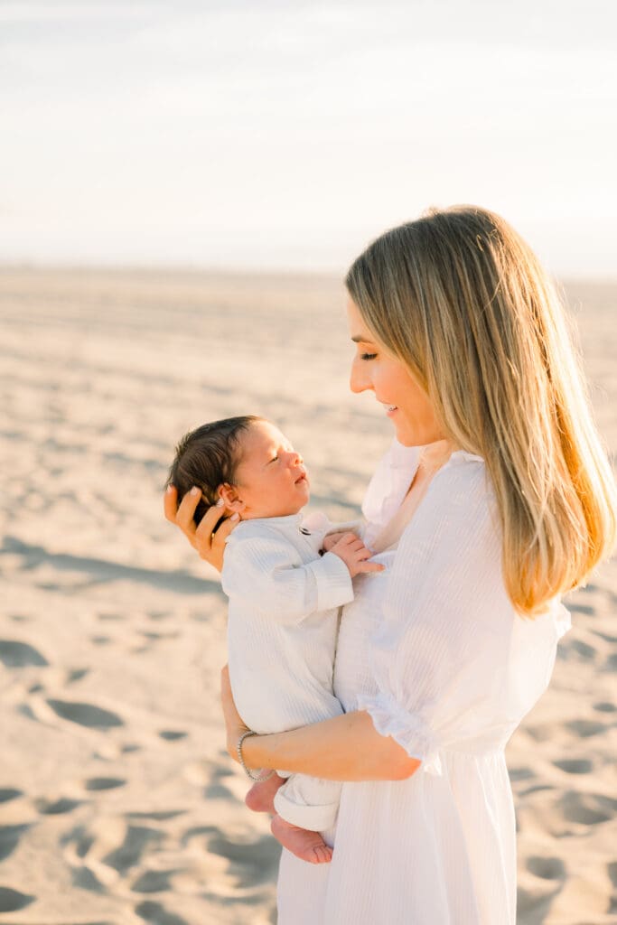 A Hermosa Beach newborn photographer takes a photo of a mom holding her newborn baby on the beach at sunset.