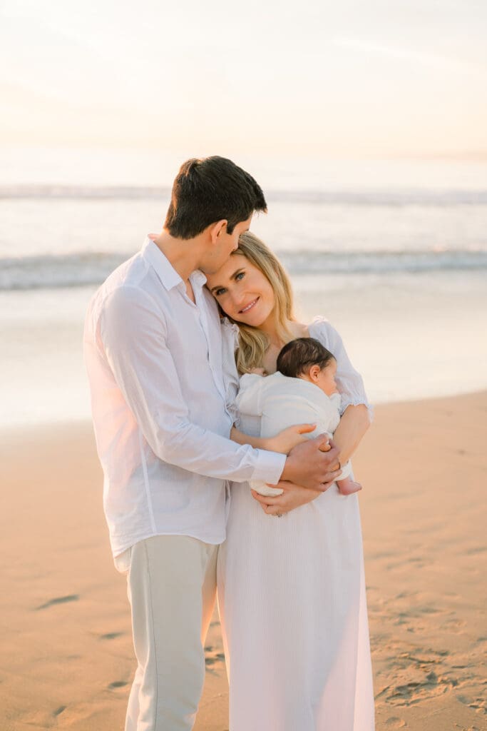 Dad kisses mom's head while she holds their newborn baby during their newborn photo shoot in Hermosa Beach.