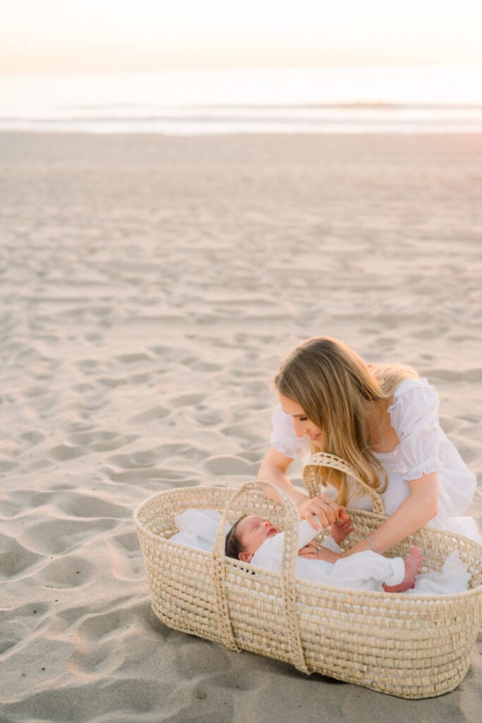Mom checks on baby in a wicker basket on the beach while taking newborn photos.