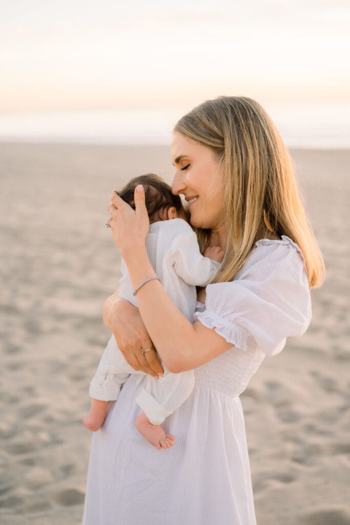 Mom holds her newborn baby on the beach while being photographed by a Hermosa Beach newborn photographer.