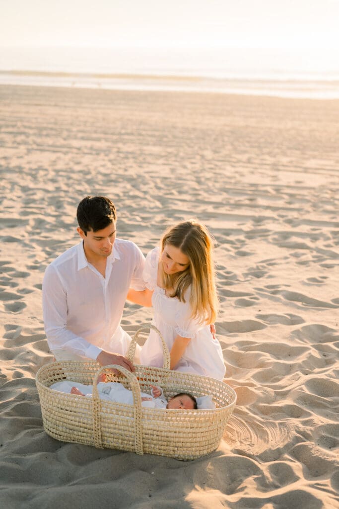 A new mom and dad look down at their baby in a wicker moses basket on the beach at sunset during their newborn photo shoot in Hermosa Beach.