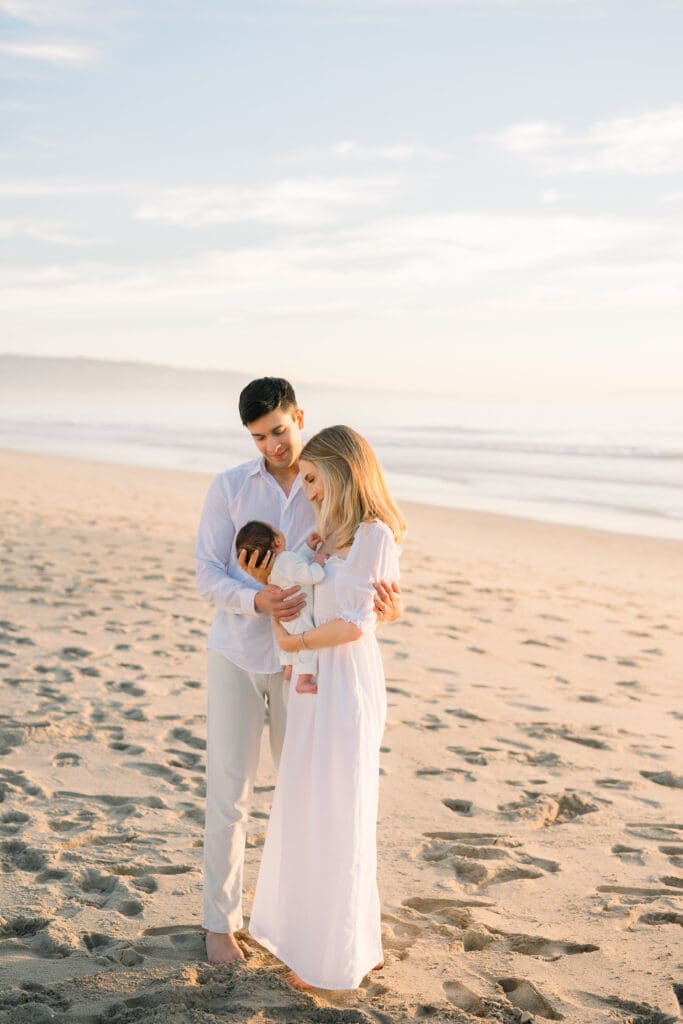 A mom and dad look lovingly at their newborn baby on the beach.