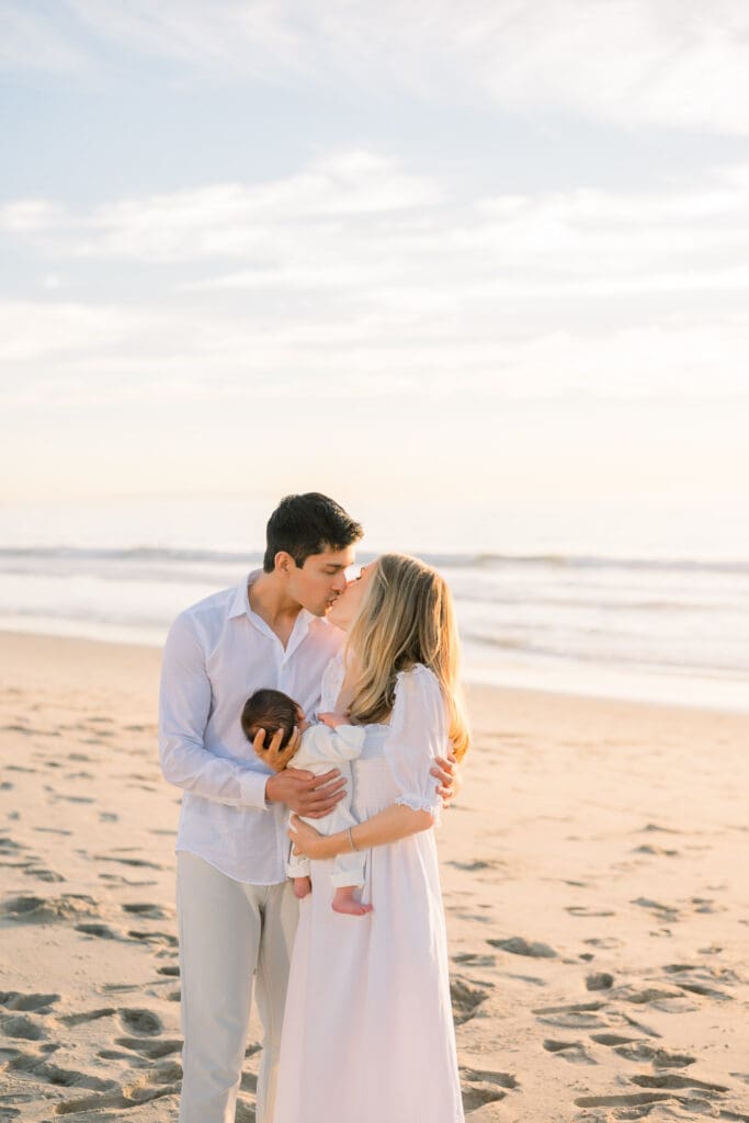 A mom and dad kiss while holding their newborn baby and being photographed by a Hermosa Beach newborn photographer.