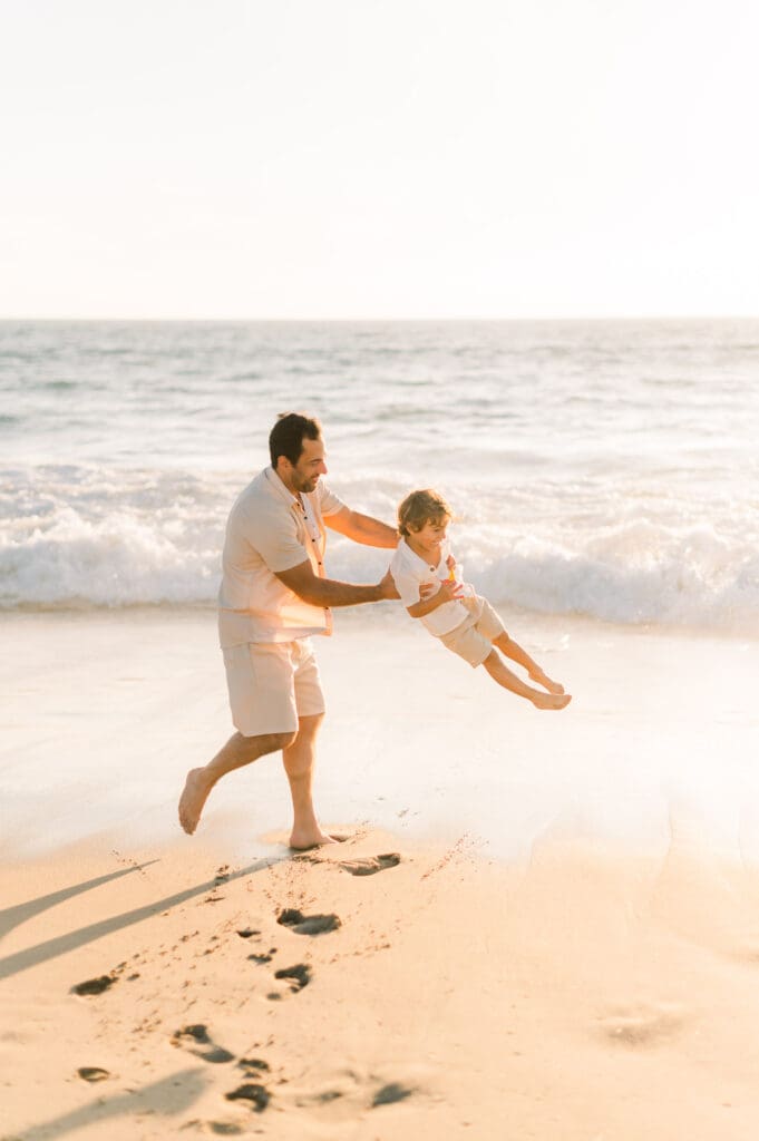 A dad spinning his son around on the beach at sunset.