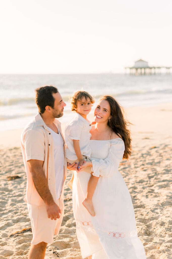 A mom, dad, and son posing for maternity photos on the beach with the Manhattan Beach Pier in the background.