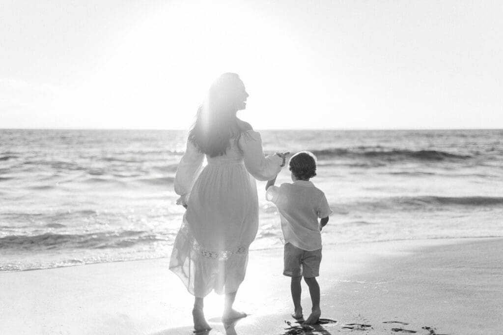 A mom and son hold hands and walk on the beach at sunset.