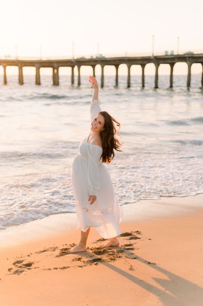 A pregnant woman in a white dress dances on the beach at sunset taking maternity photos.