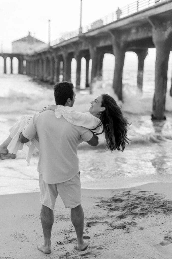 A man carries his pregnant wife in front of the Manhattan Beach pier during their maternity photoshoot.