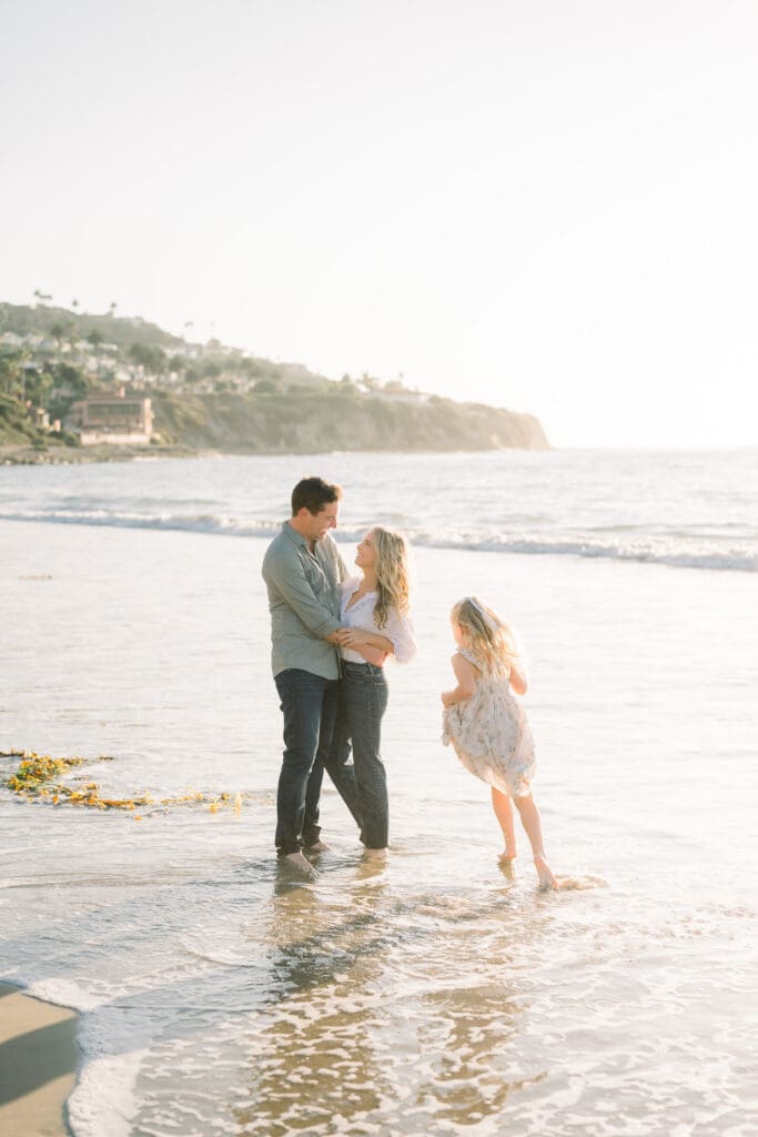 Mom and dad hugging in the ocean while their daughter runs around at sunset.