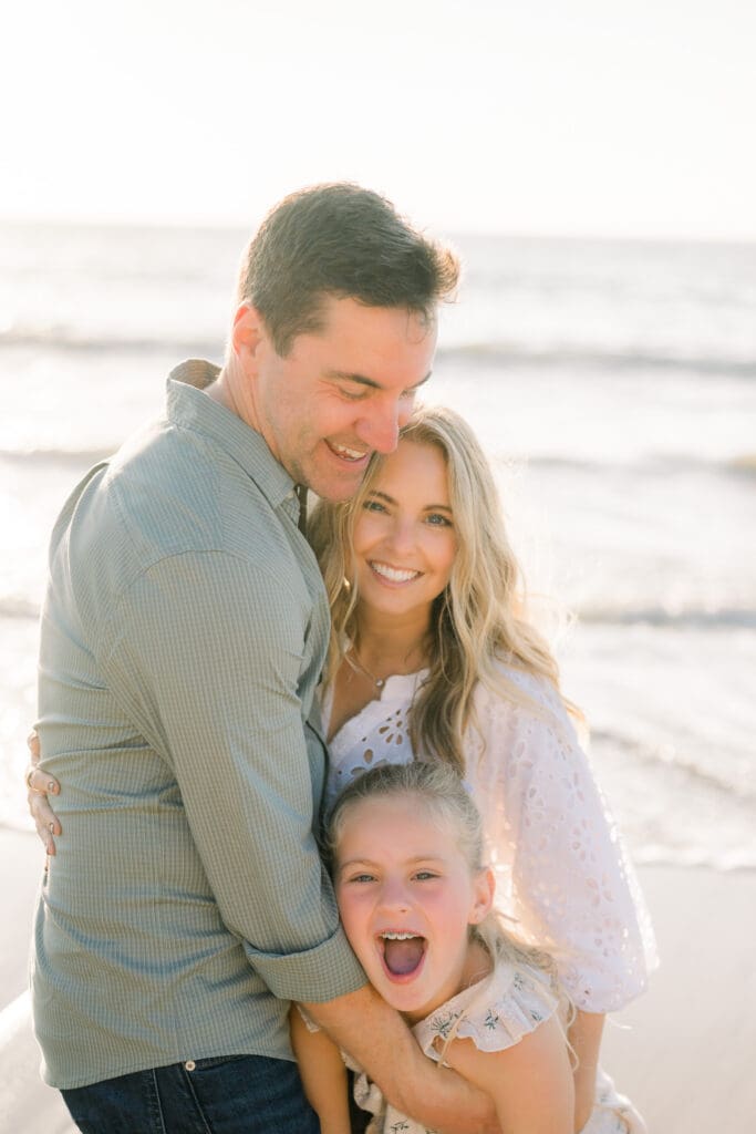 A family of three snuggled together smiling and laughing on the beach during a family photography session in Los Angeles.