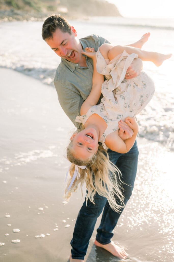 A dad playfully holding his daughter upside down while they laugh on the beach.