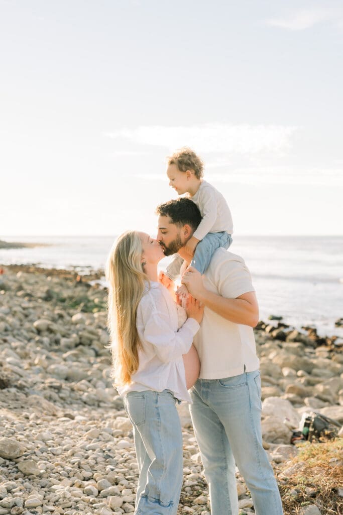 A mom and dad kissing while dad puts the toddler on his shoulders.  They are wearing jeans and white shirts for their maternity photos in Los Angeles.