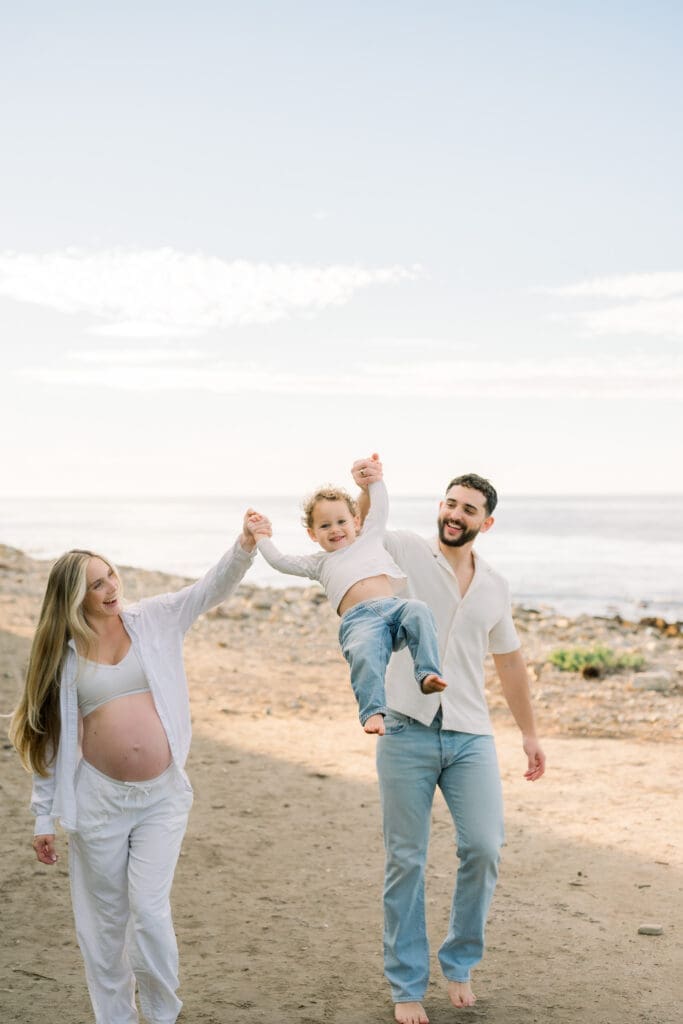 A family playing on the beach while taking maternity photos and wearing jeans and white tops.
