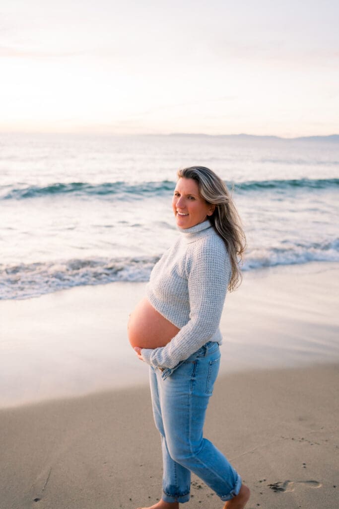 A woman taking maternity photos while wearing jeans and sweater on the beach.