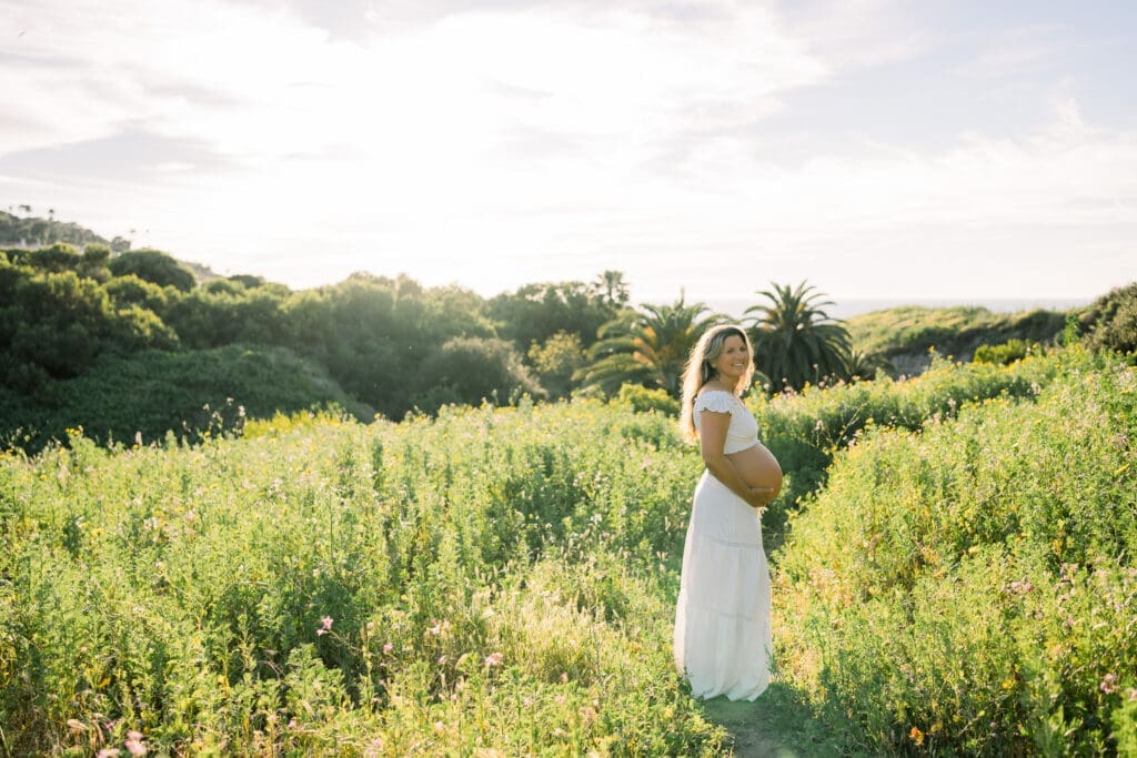 A mama to be posing for maternity photos in a flower field wearing a white 2 piece skirt and top that shows her belly.