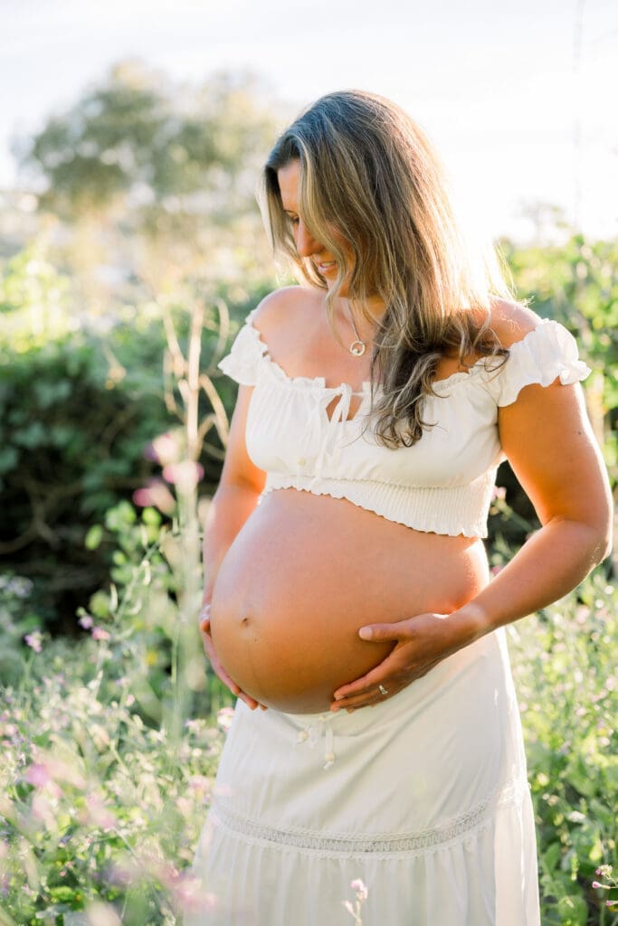 A mama to be holding her belly wearing a white skirt and off the shoulder top while posing for maternity photos in Los Angeles.