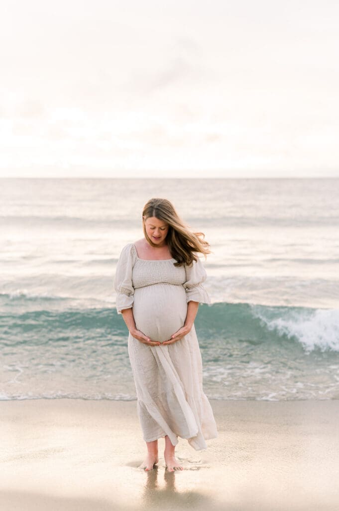 A pregnant woman on the beach wearing a flowy cream dress.
