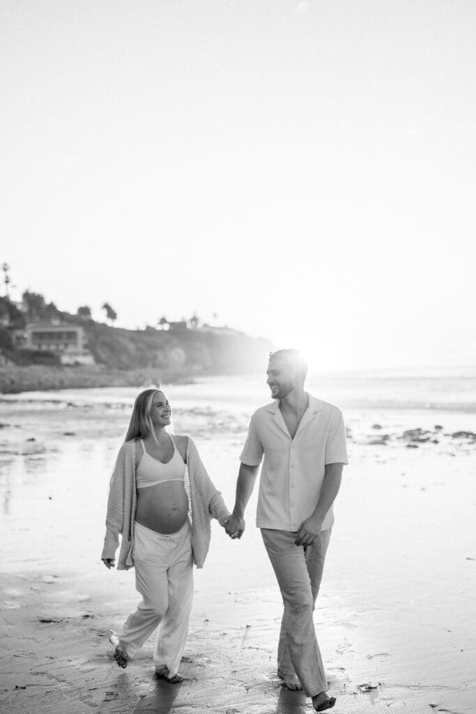 A black and white photo of a couple walking on the beach while taking maternity photos.