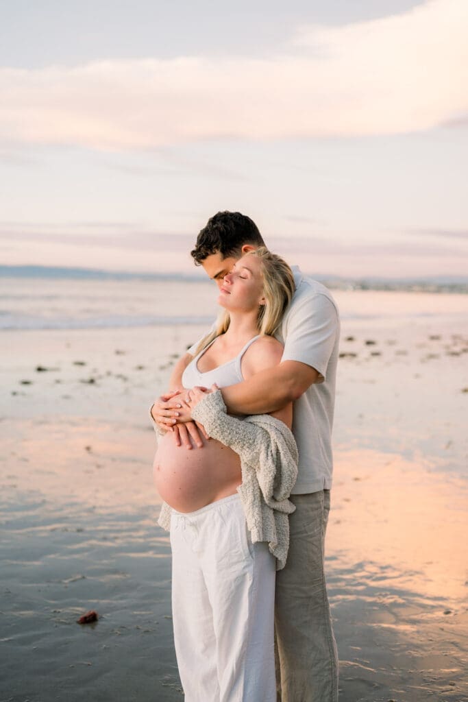 A couple embracing on the beach in a sweet maternity photo pose.