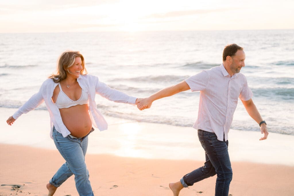A couple holding hands and running on the beach during their Los Angeles maternity session.
