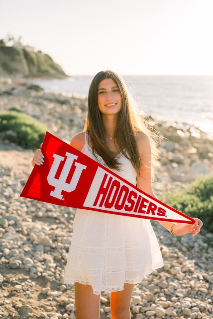 A Los Angeles senior photo session featuring a girl in a white dress posing on the beach with her future college banner.
