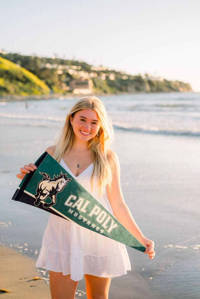 A senior girl holds a college banner as she smiles on the beach.